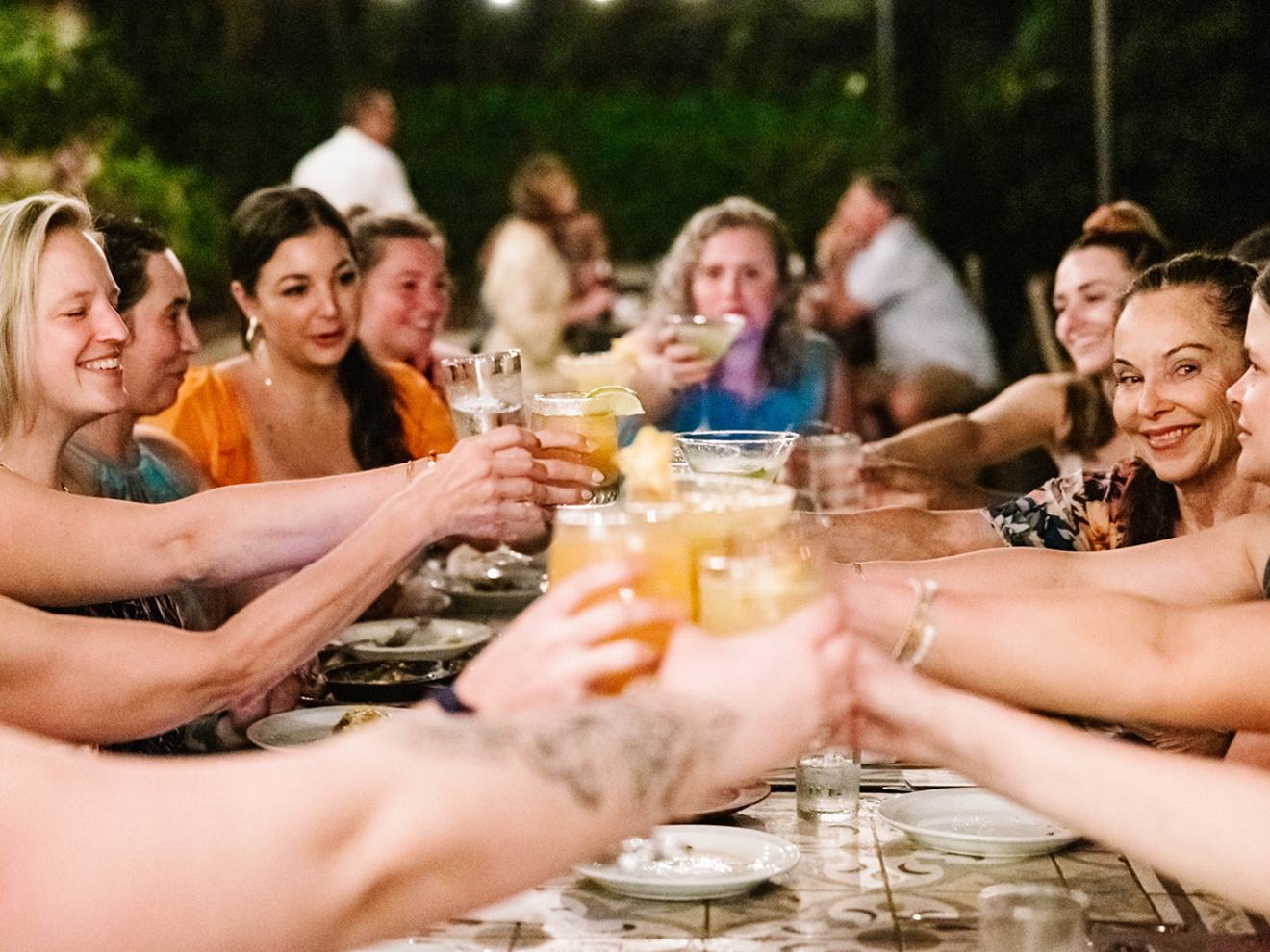 Group of people toasting with drinks at a long table outdoors.