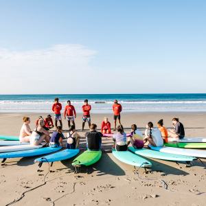 Group in surf lesson on beach, seated on surfboards, instructors in red, ocean in background.