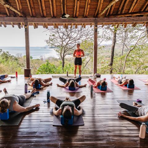 People doing yoga on mats with a view of the ocean under a wooden pavilion.