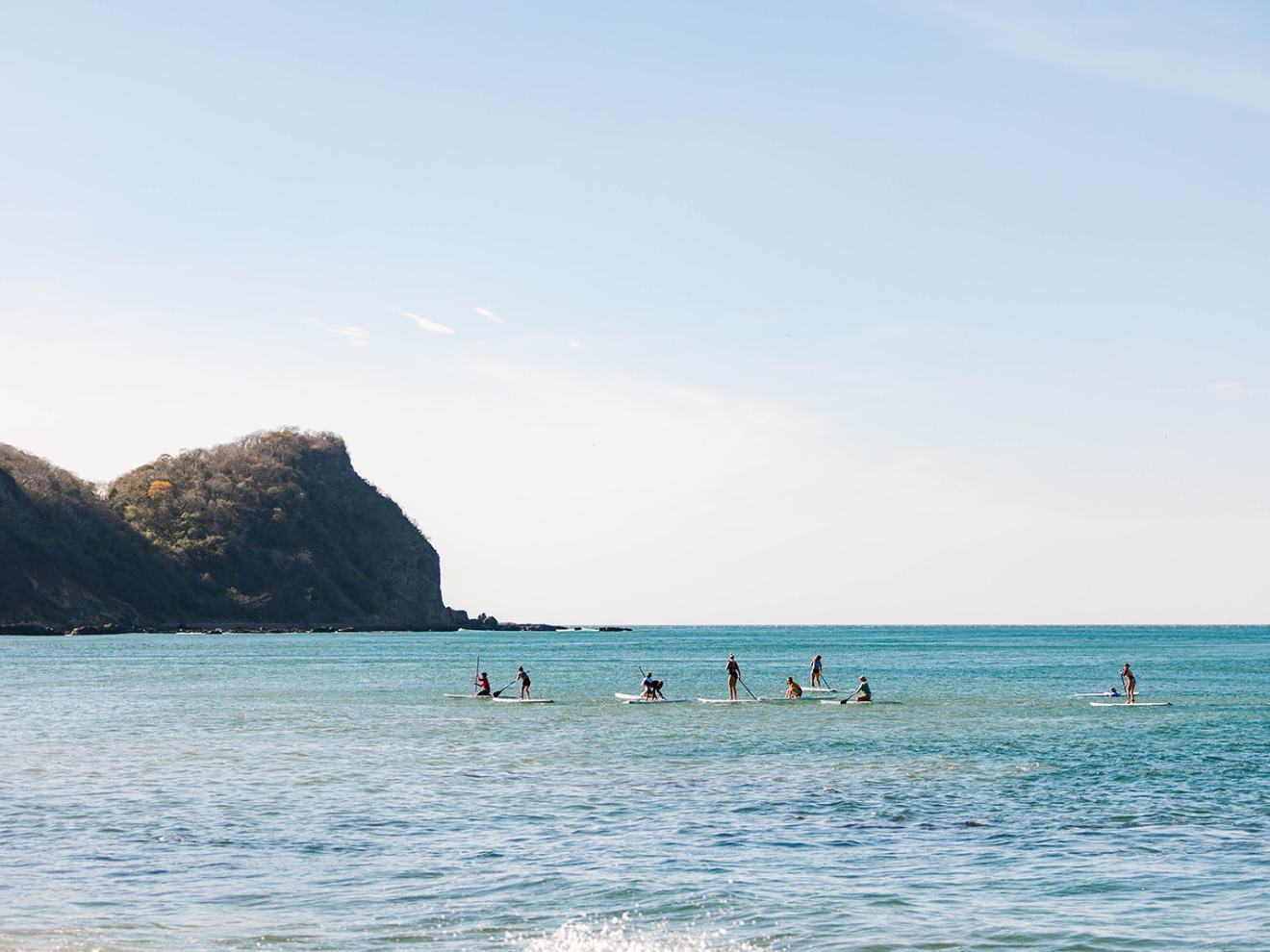 People paddleboarding on calm sea near rocky coastline under blue sky.
