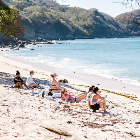 People sitting on a sandy beach with hills and ocean in the background.