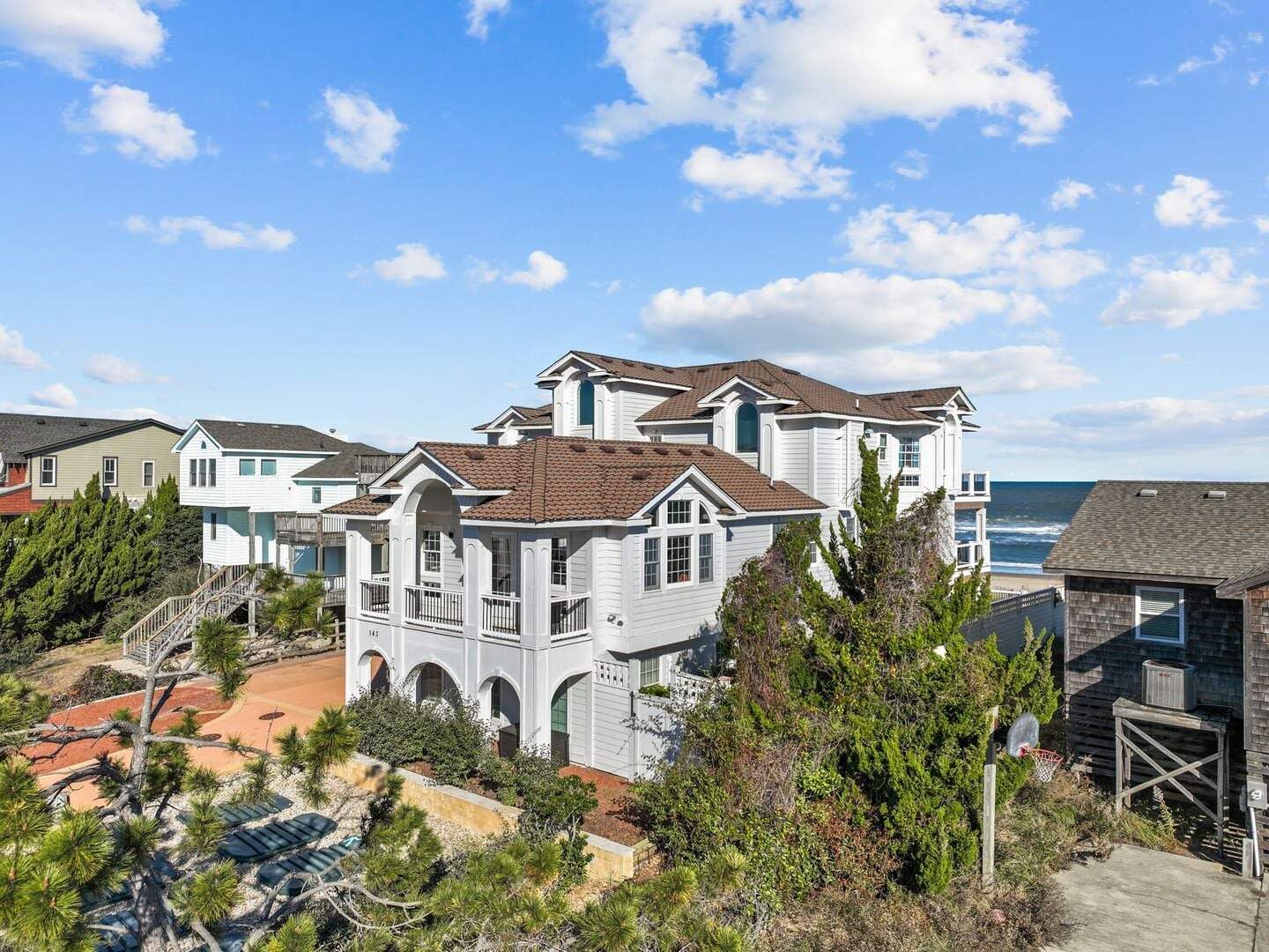 A large multi-story house with a brown roof overlooks the ocean, surrounded by other homes and greenery.