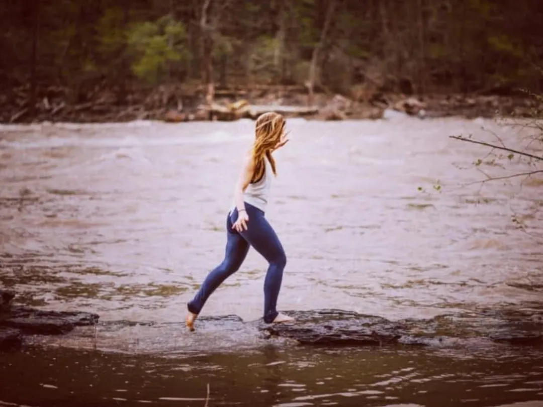 Person in leggings stepping on wet rocks in a river, surrounded by forest.