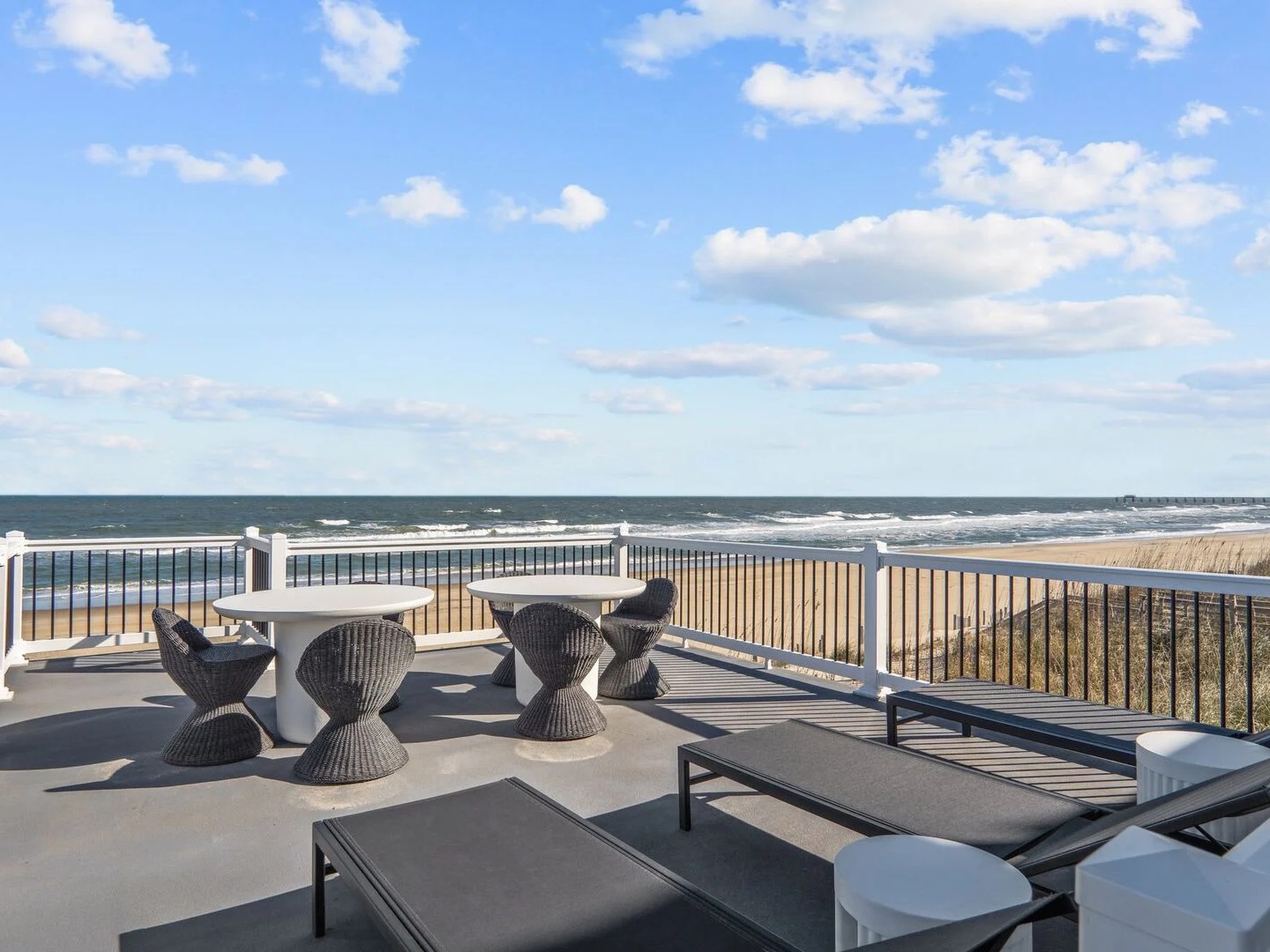 Oceanfront patio with tables, chairs, and lounge chairs overlooking a sandy beach and blue sky.