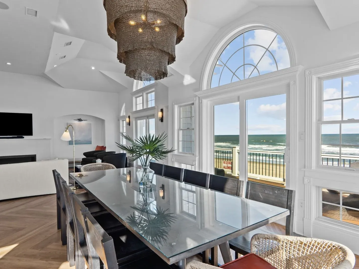 Dining room with glass table, wicker chairs, and ocean view through large windows.