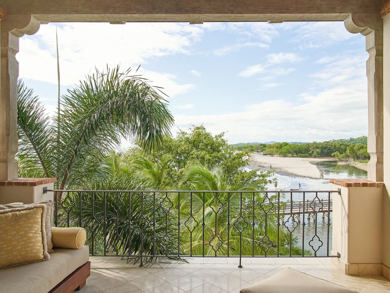 Balcony view of tropical trees, sandy beach, and ocean under a partly cloudy sky.