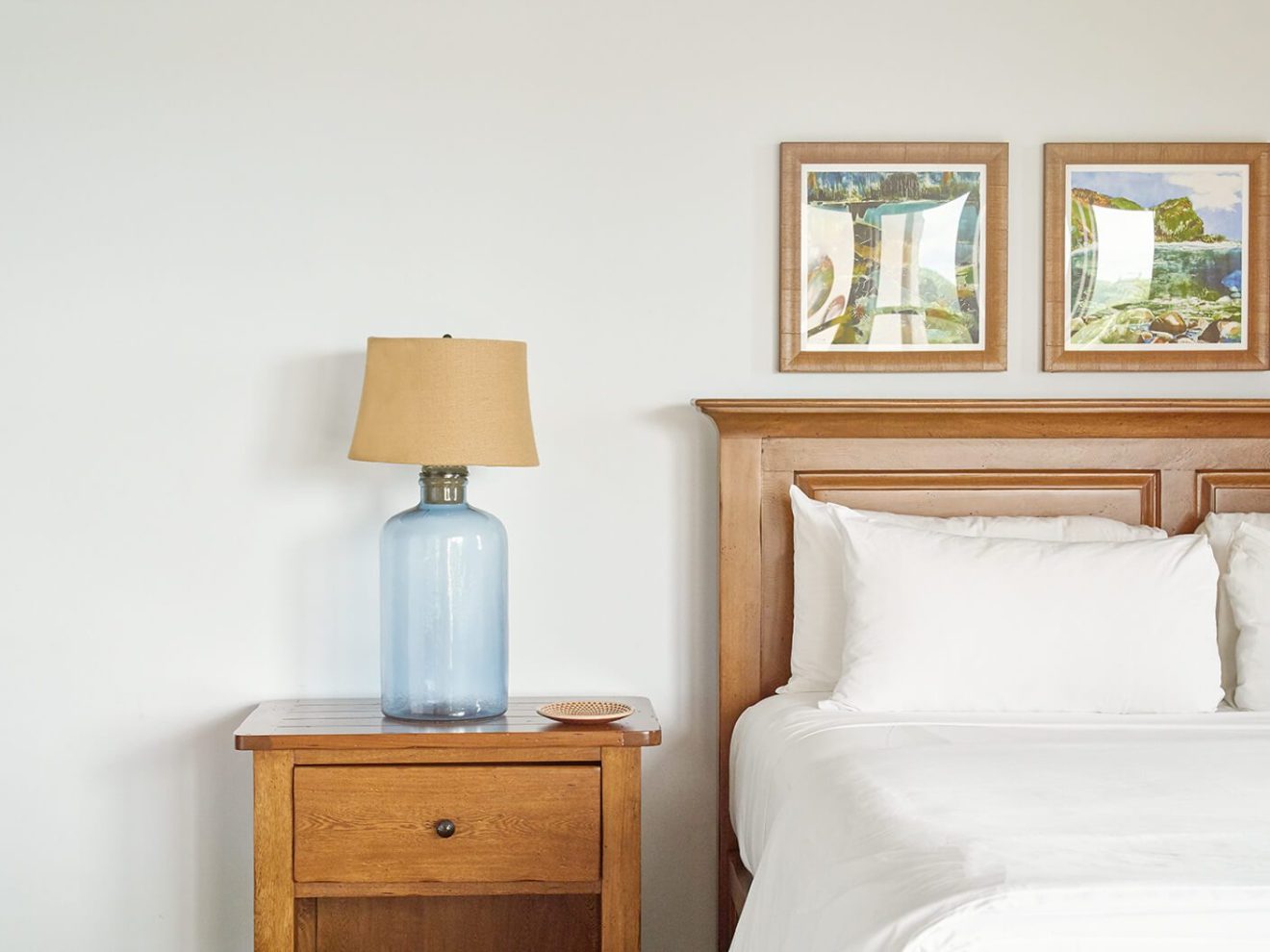Wooden bed and nightstand with lamp, framed artwork on white wall.