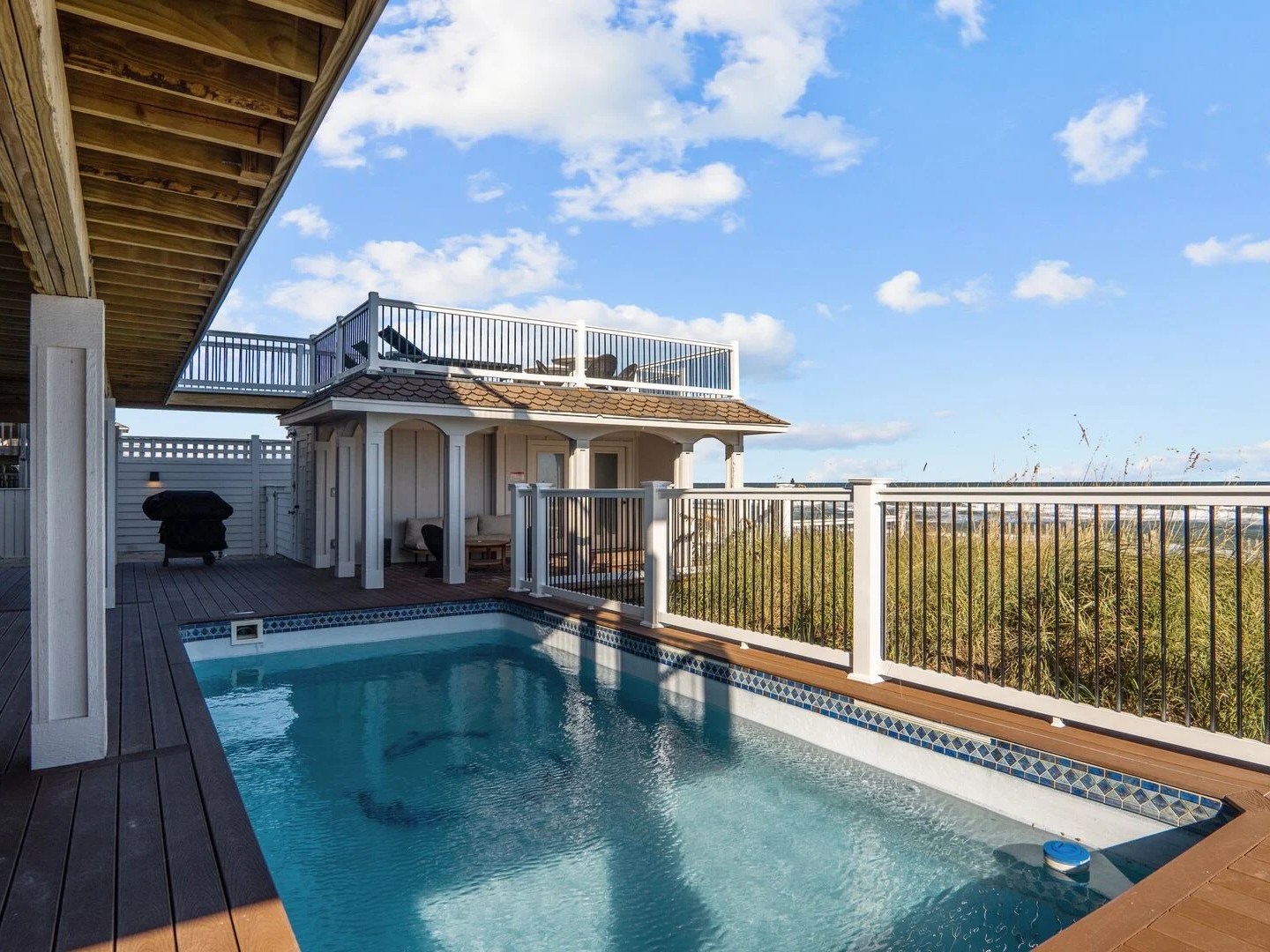 Outdoor pool with a wooden deck and a two-story pavilion under a blue sky with clouds.
