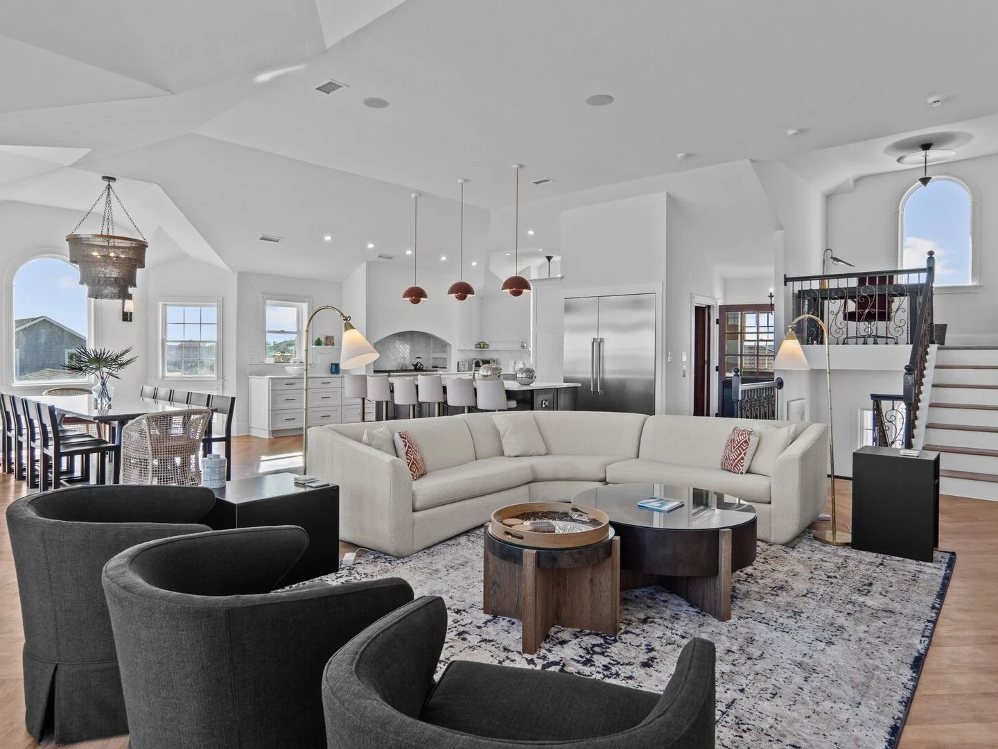 Modern open-plan living room with beige sofa, dark chairs, dining area, kitchen, and stairs.