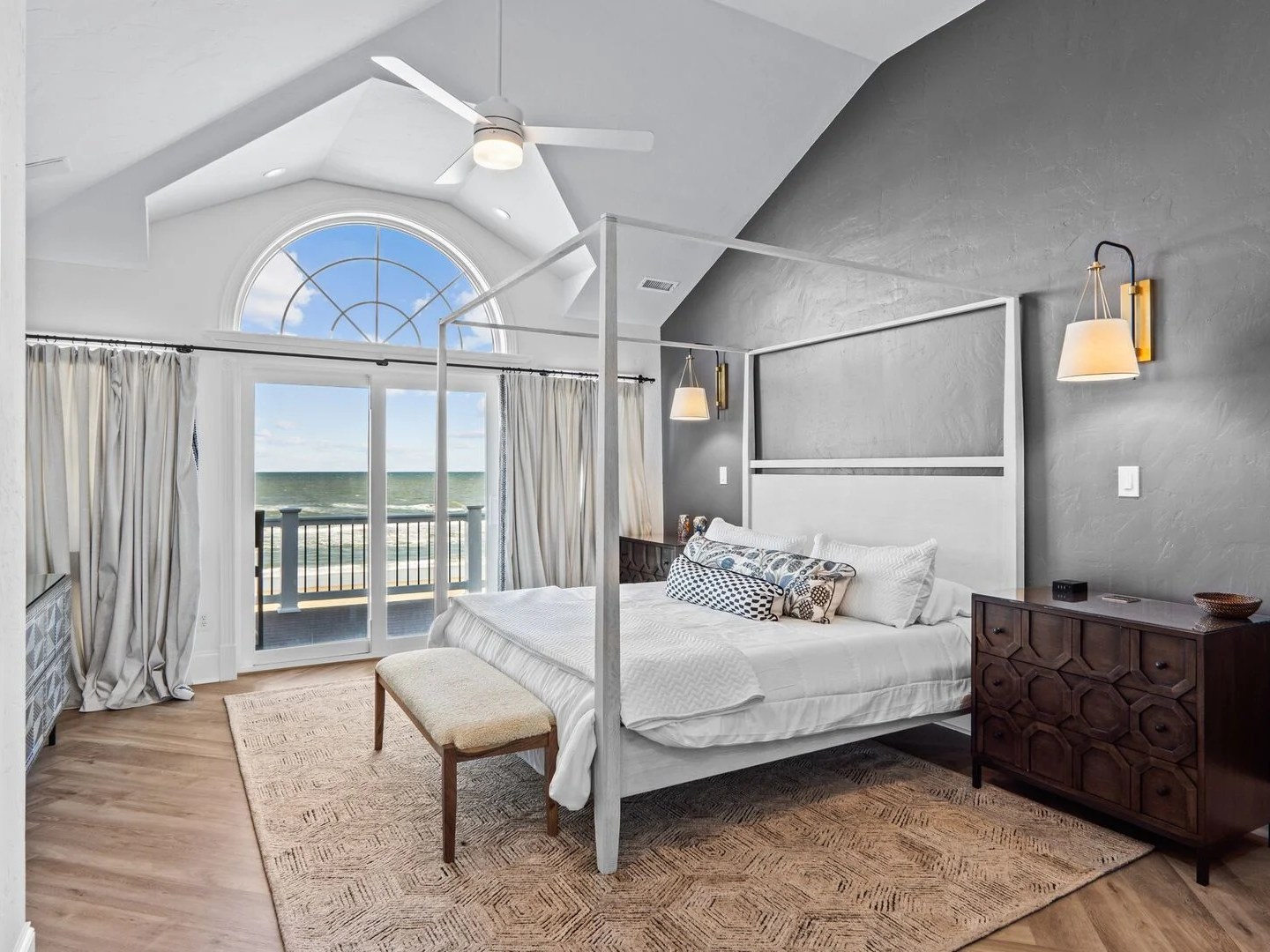 Coastal bedroom with gray walls, canopy bed, and ocean view through arched window.