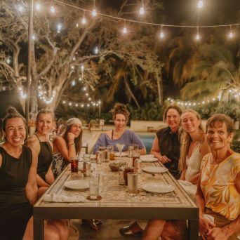Seven people smiling at a table outdoors, under string lights at night.