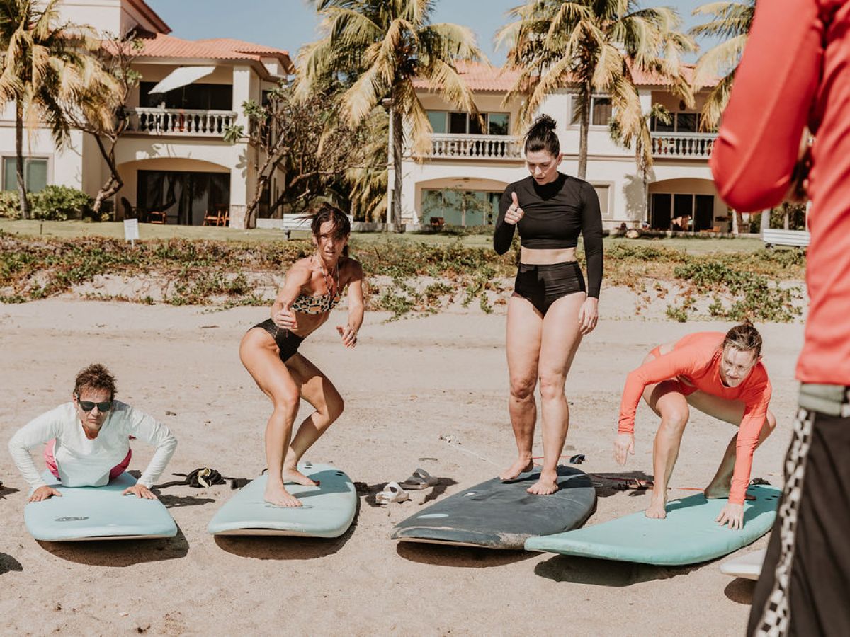 Four people practicing surfing stances on a beach, with palm trees and houses in the background.