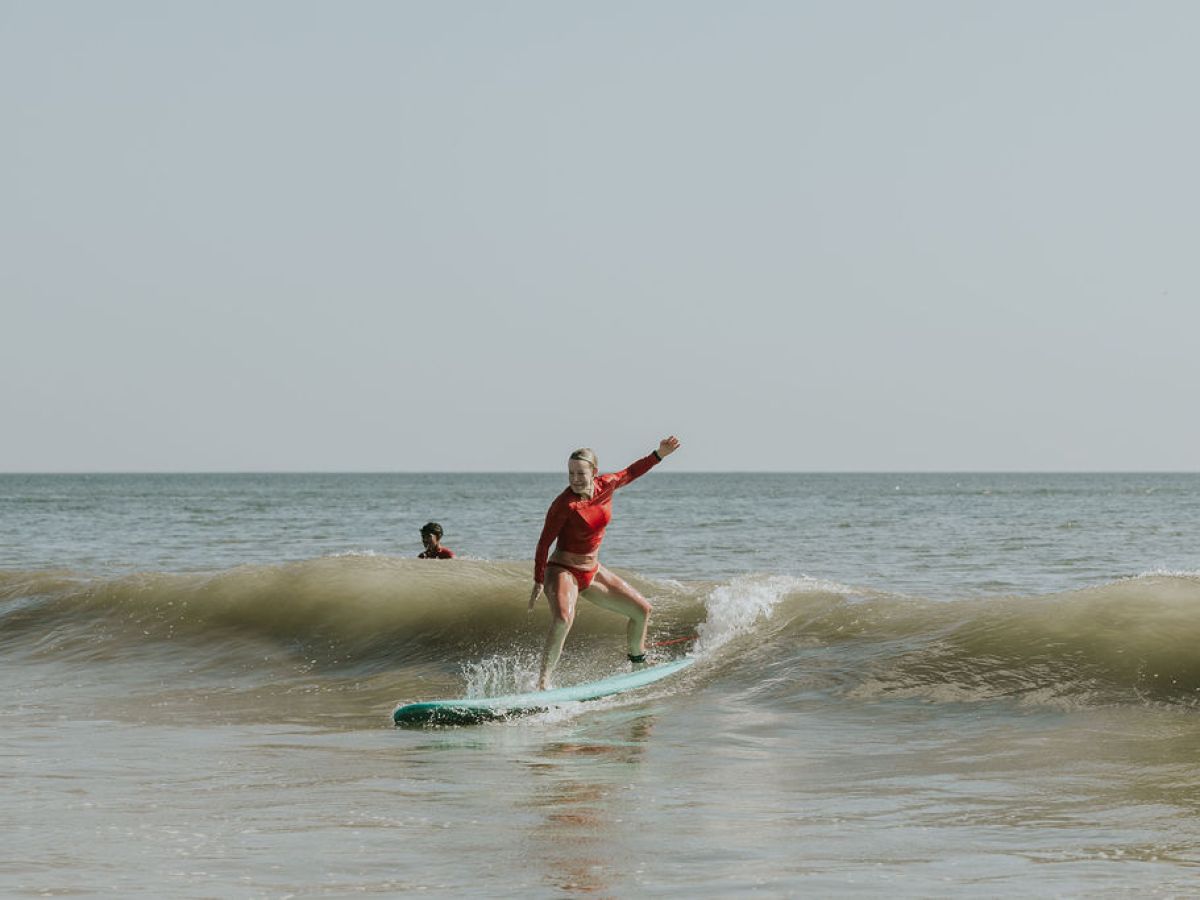 Person in red surfs a wave, another person watches in the background.