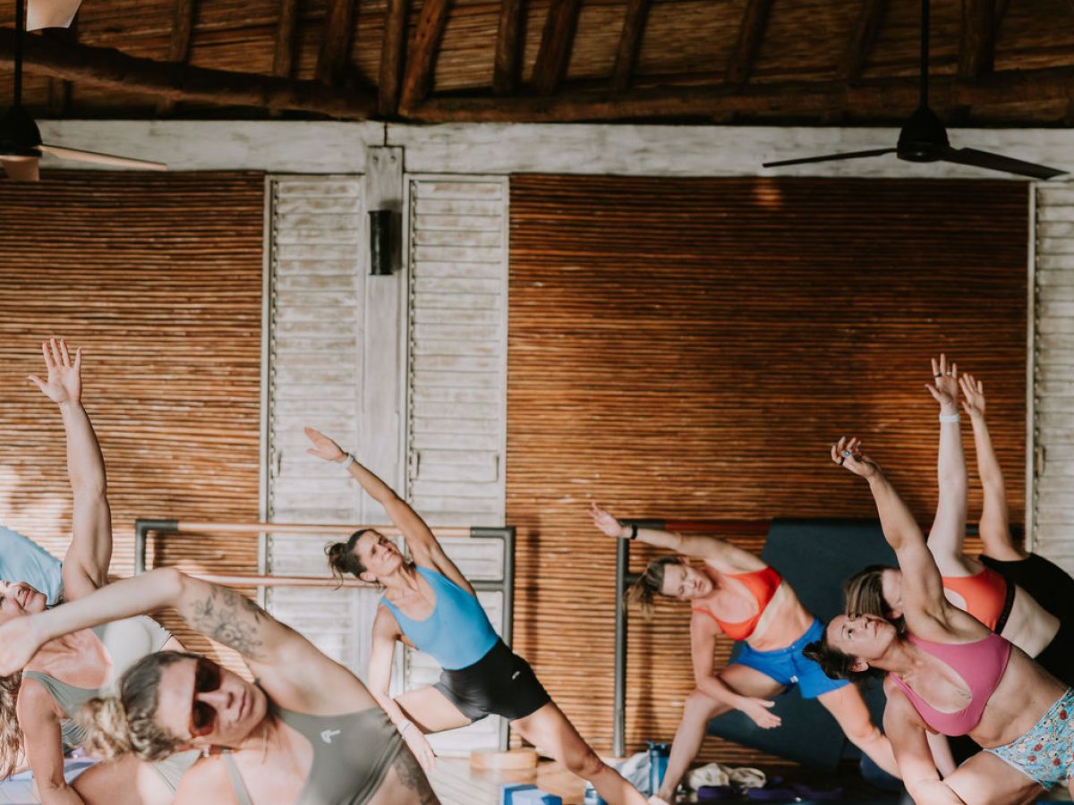 Group of people doing yoga in a wooden studio, performing side stretches with raised arms.