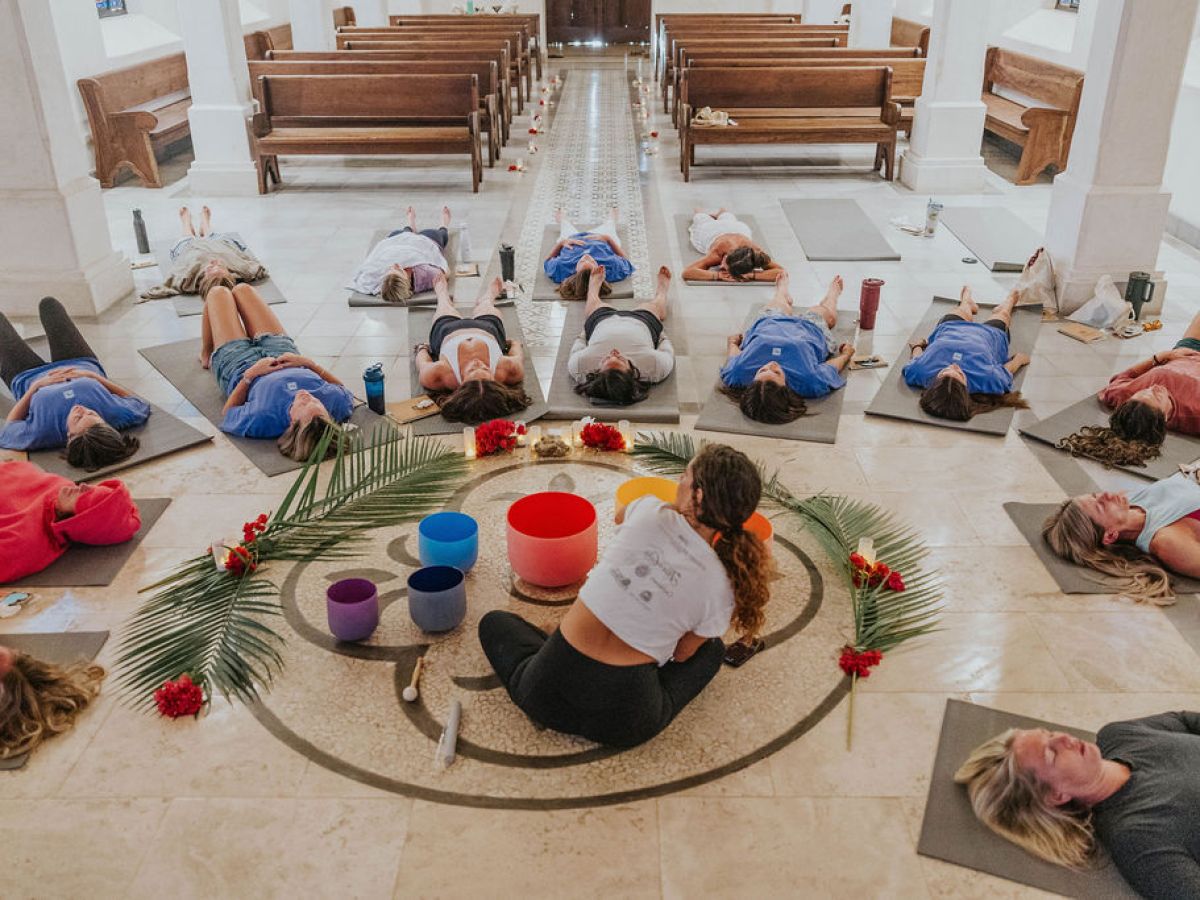 Group lying on yoga mats in a circle with colorful bowls and palm leaves in a chapel.