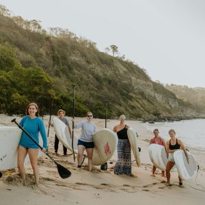 Six women with paddleboards and paddles on a sandy beach, with cliffs and ocean in the background.