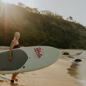 Person holding a paddleboard on a sunny beach with cliffs in the background.