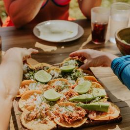 Tacos with lime on a wooden platter, hands reaching, and a person holding a phone at a table.
