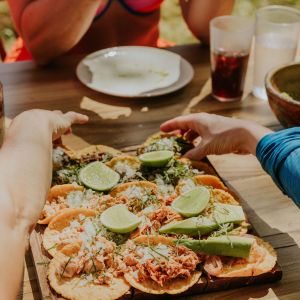 Tacos with lime on a wooden platter, hands reaching, and a person holding a phone at a table.
