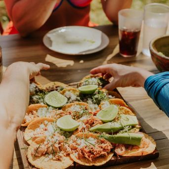 Tacos with lime on a wooden platter, hands reaching, and a person holding a phone at a table.