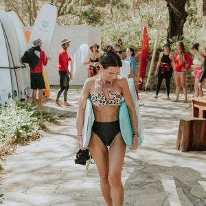 Woman in swimsuit carrying surfboards, surrounded by people and trees.