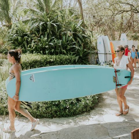 Two women carrying a blue surfboard on a sunny path surrounded by greenery.