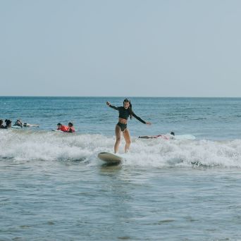 Person surfing on a wave with others in the background on a sunny beach day.