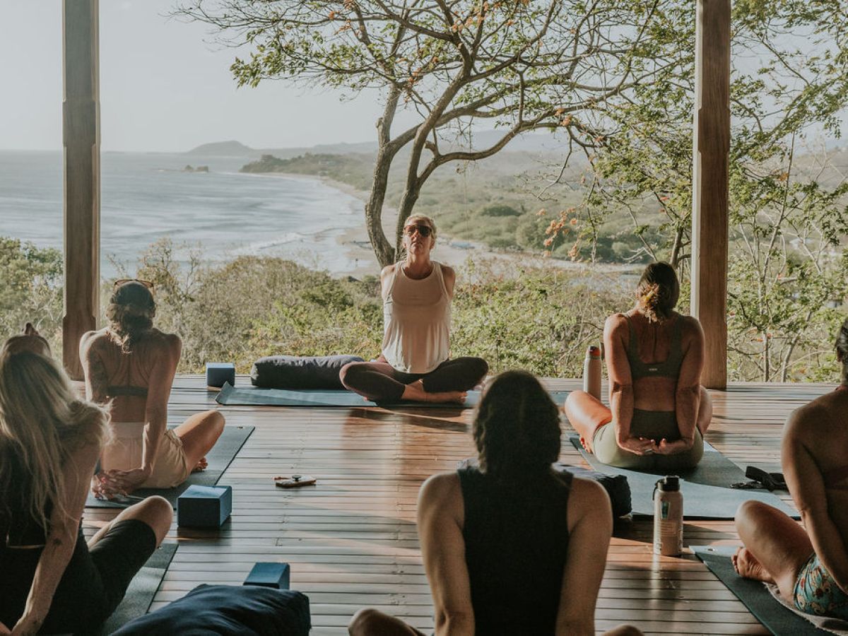 Group yoga session on wooden deck with ocean view and trees in the background.