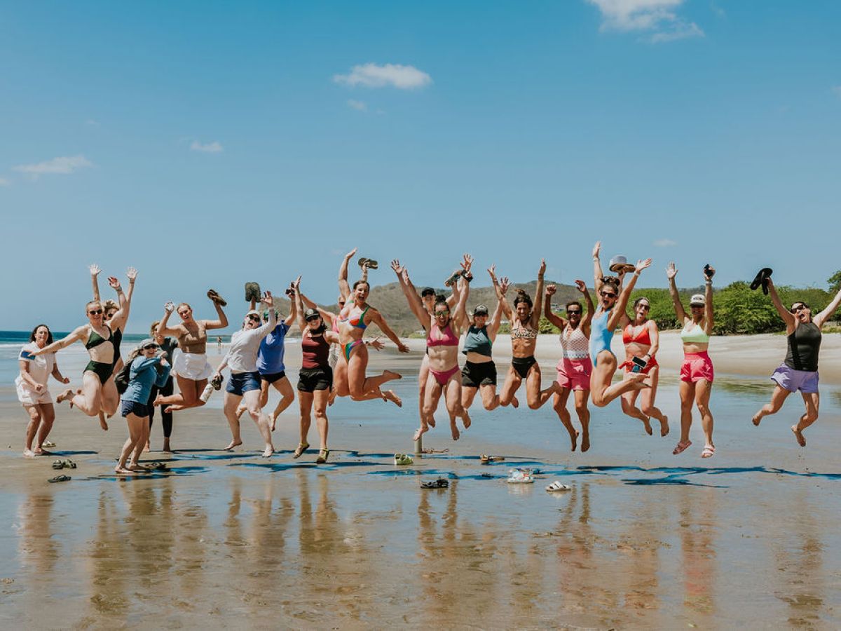 Group of people jumping on a sunlit beach against a blue sky.