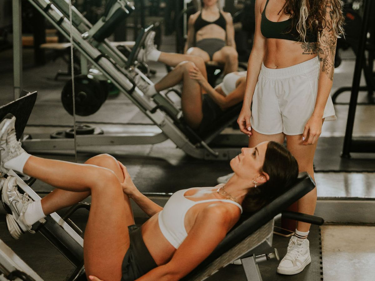 Woman exercising on leg press machine with another woman standing beside her in a gym.