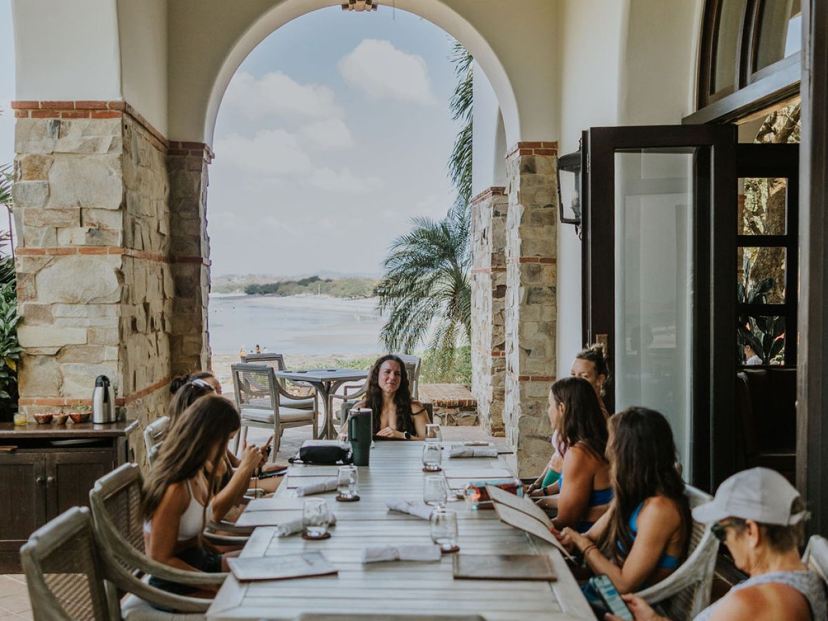 Group of people sitting at an outdoor dining table under an archway with a view of the beach.