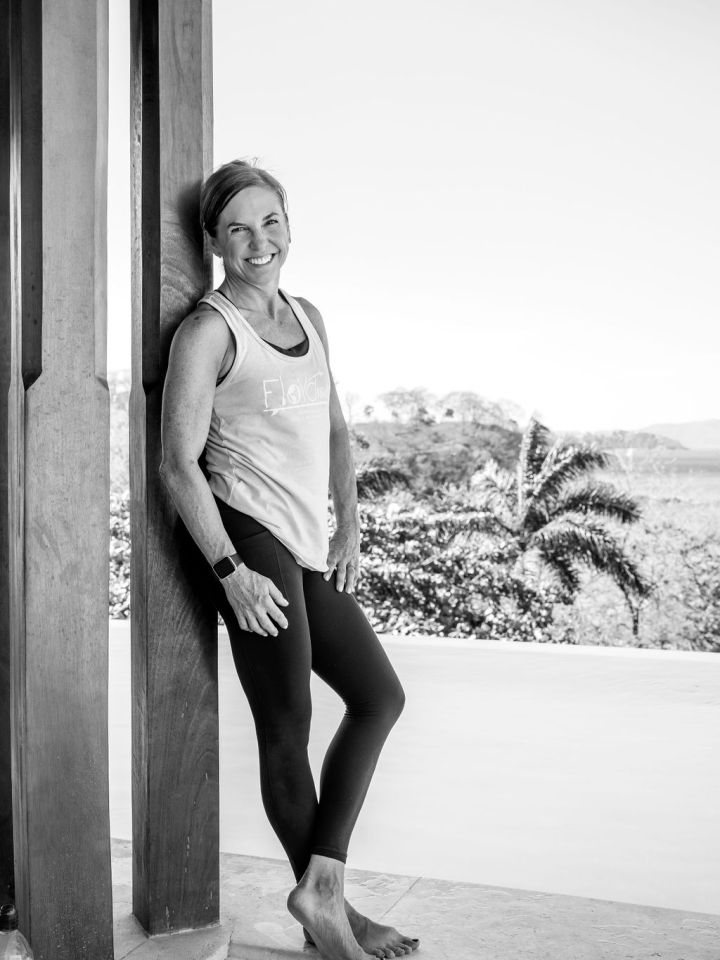 Woman in activewear smiling, leaning against a wooden post outdoors.