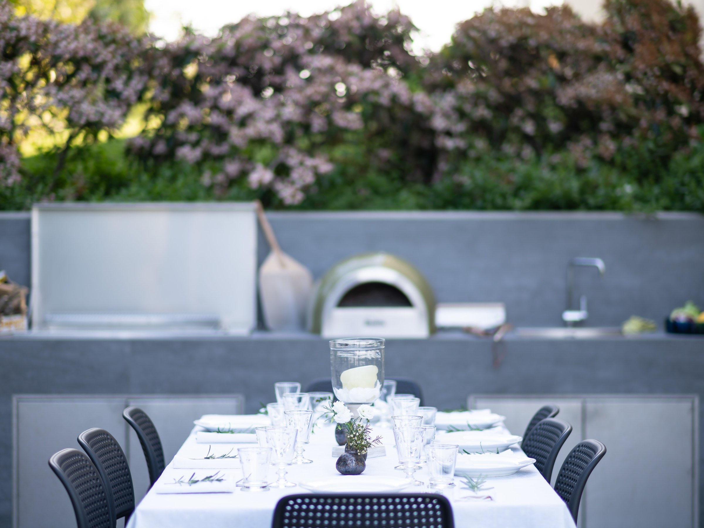 Outdoor dining table set with white cloth, glassware, near an outdoor kitchen with a pizza oven.