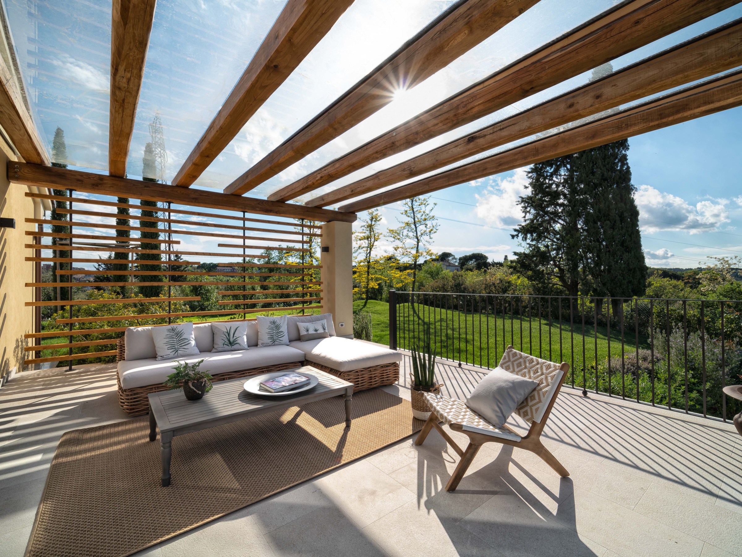 Outdoor patio with wooden pergola, white sofa, chairs, table, and view of garden under blue sky.