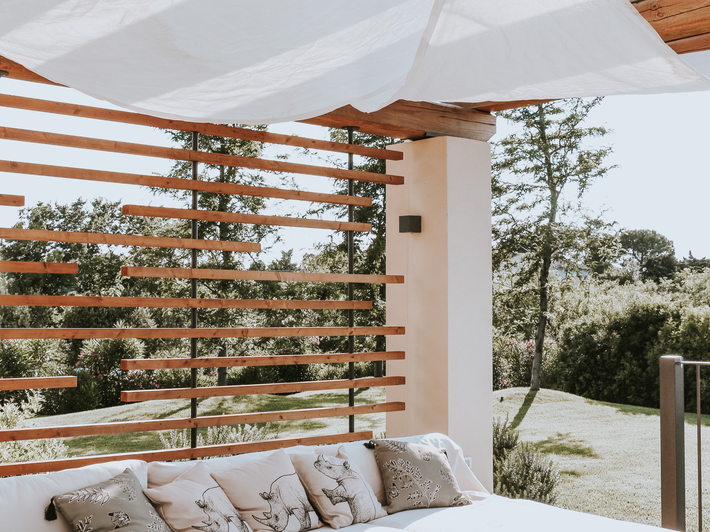 Outdoor patio with wicker sofa, white cushions, fabric canopy, and wooden slat walls in sunny garden setting.