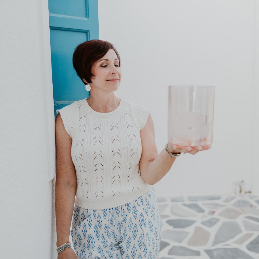 Woman with short hair holding a transparent object, standing by a blue window on a stone-paved floor.