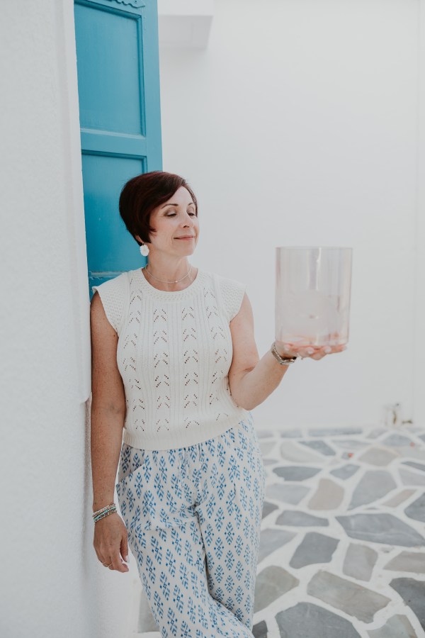 Woman with short hair holding a transparent object, standing by a blue window on a stone-paved floor.