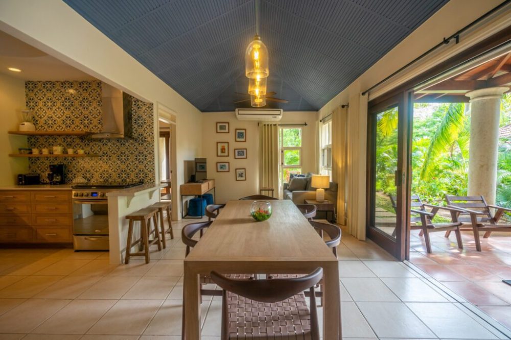 Open-plan room with dining table, kitchen, and living area, opening to a patio with greenery.