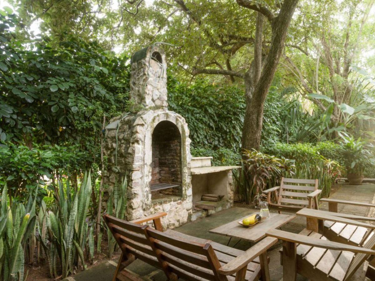 Outdoor patio with wooden furniture and a stone barbecue surrounded by lush greenery.