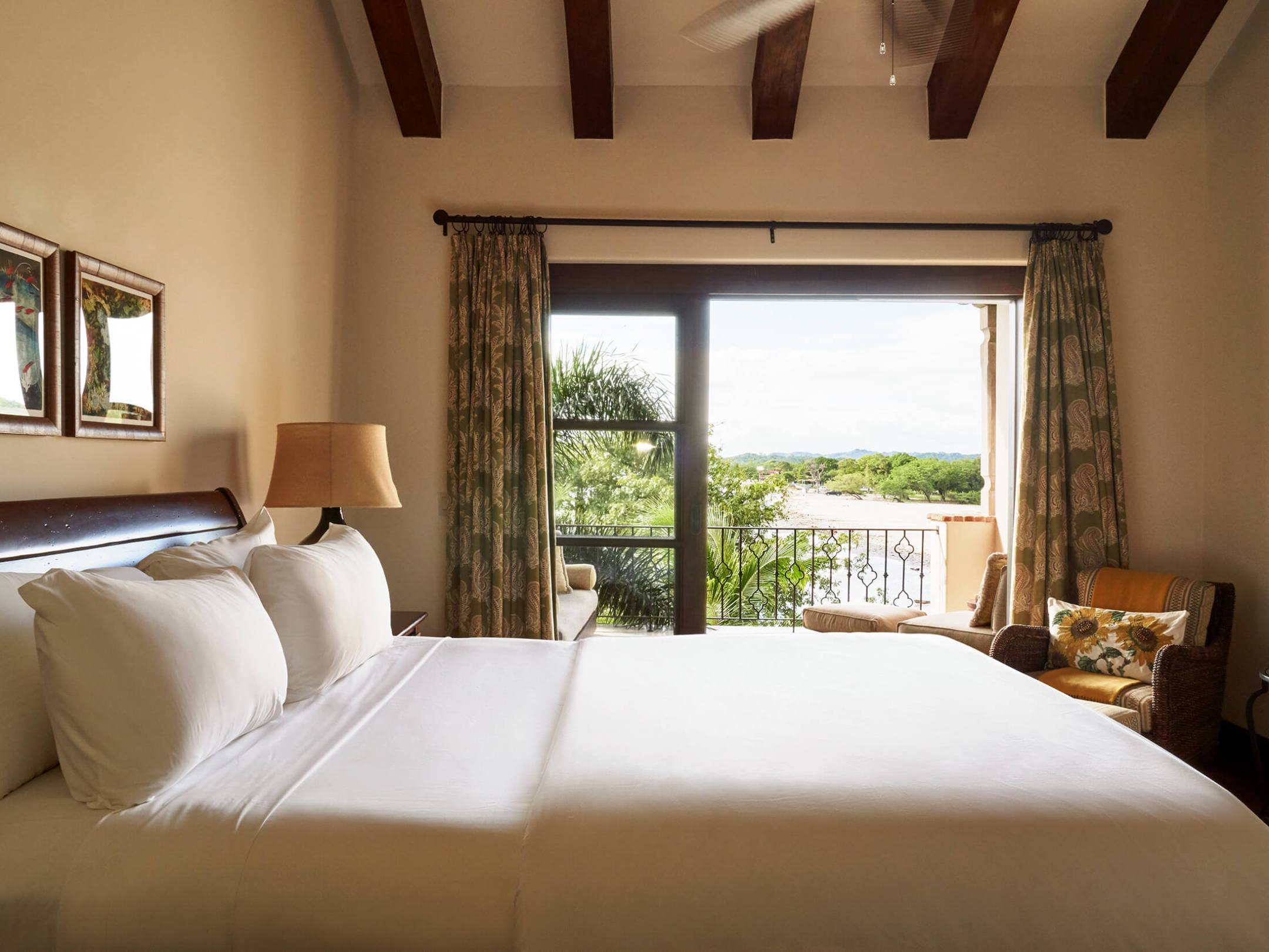 Cozy bedroom with white bedding, balcony view, wooden beams, and a lamp.