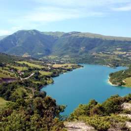Scenic view of a blue lake surrounded by green hills and mountains under a clear sky.