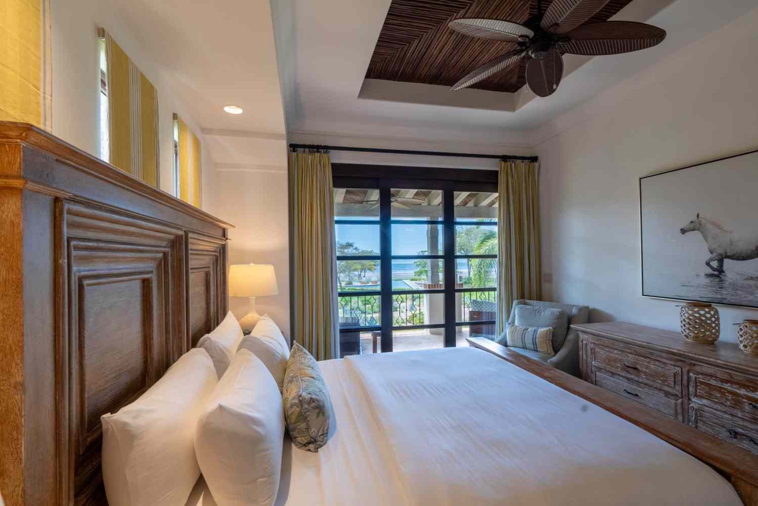 Bedroom with wooden bed, ceiling fan, and a view of a balcony overlooking the ocean.