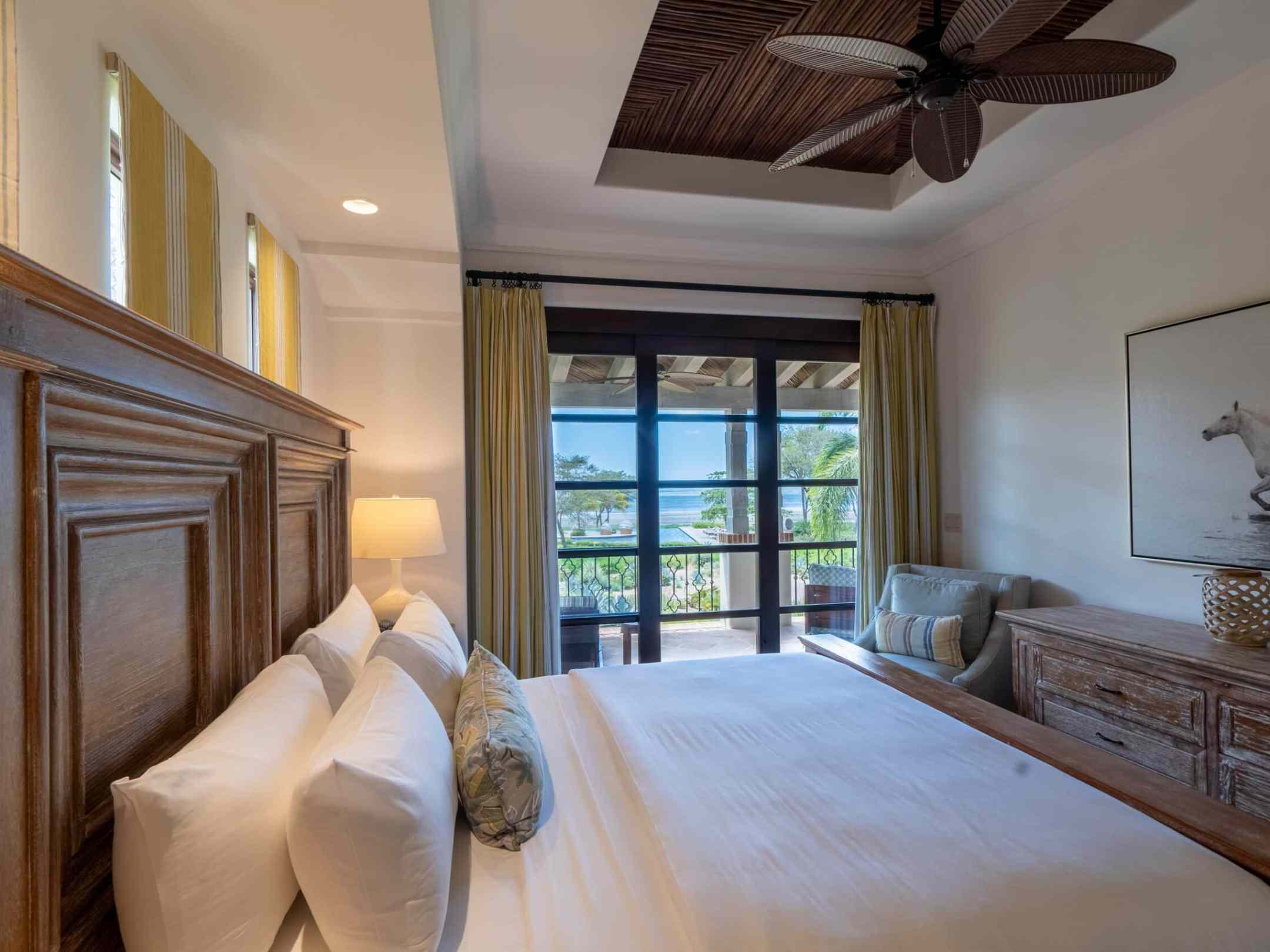 Bedroom with wooden bed, ceiling fan, and a view of a balcony overlooking the ocean.