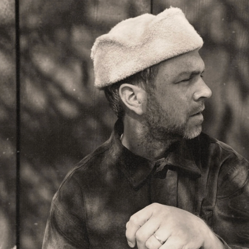 Man wearing a beret and jacket, seated outdoors.