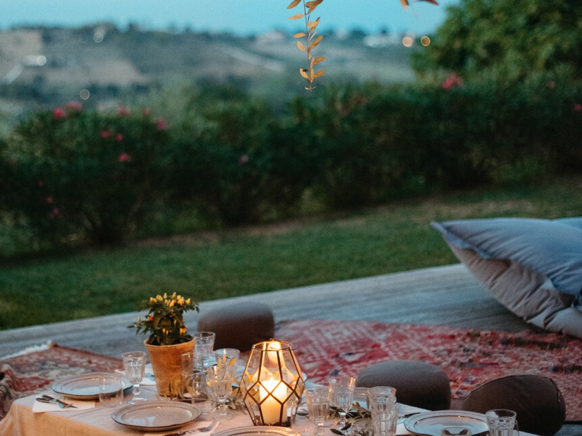 Outdoor dining setup with plates, glasses, a lit candle, and cushions under a tree at dusk.