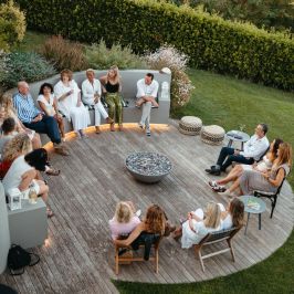 People seated in a circular outdoor gathering on wooden deck.