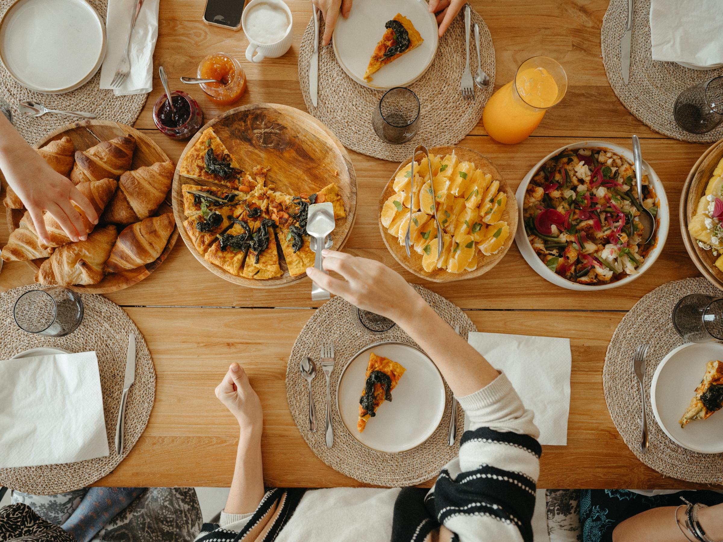 People share a meal with pizza, salad, pastries, and drinks on a wooden table.