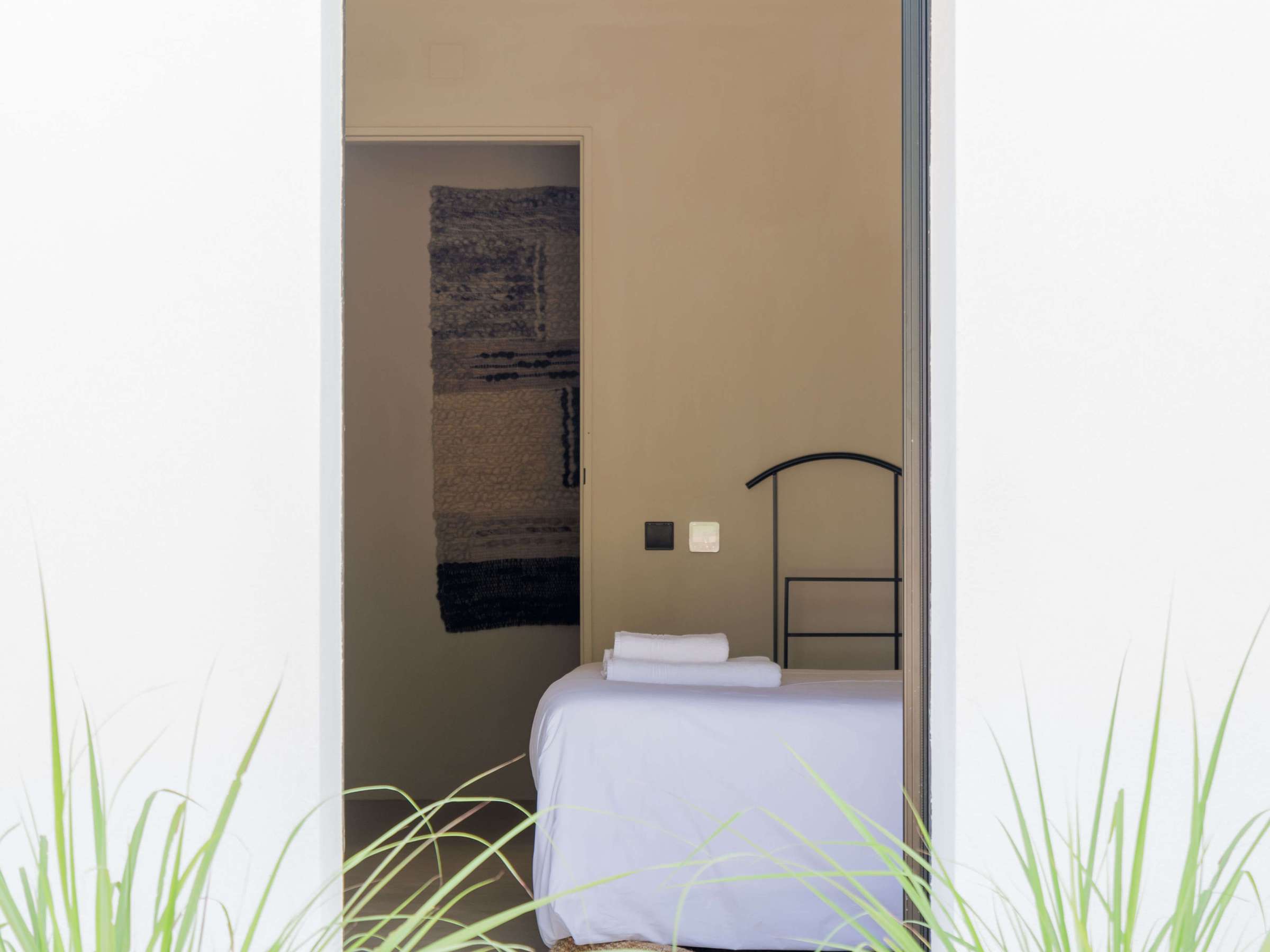 Open door showing a minimal bedroom with a made bed, framed by green grass.