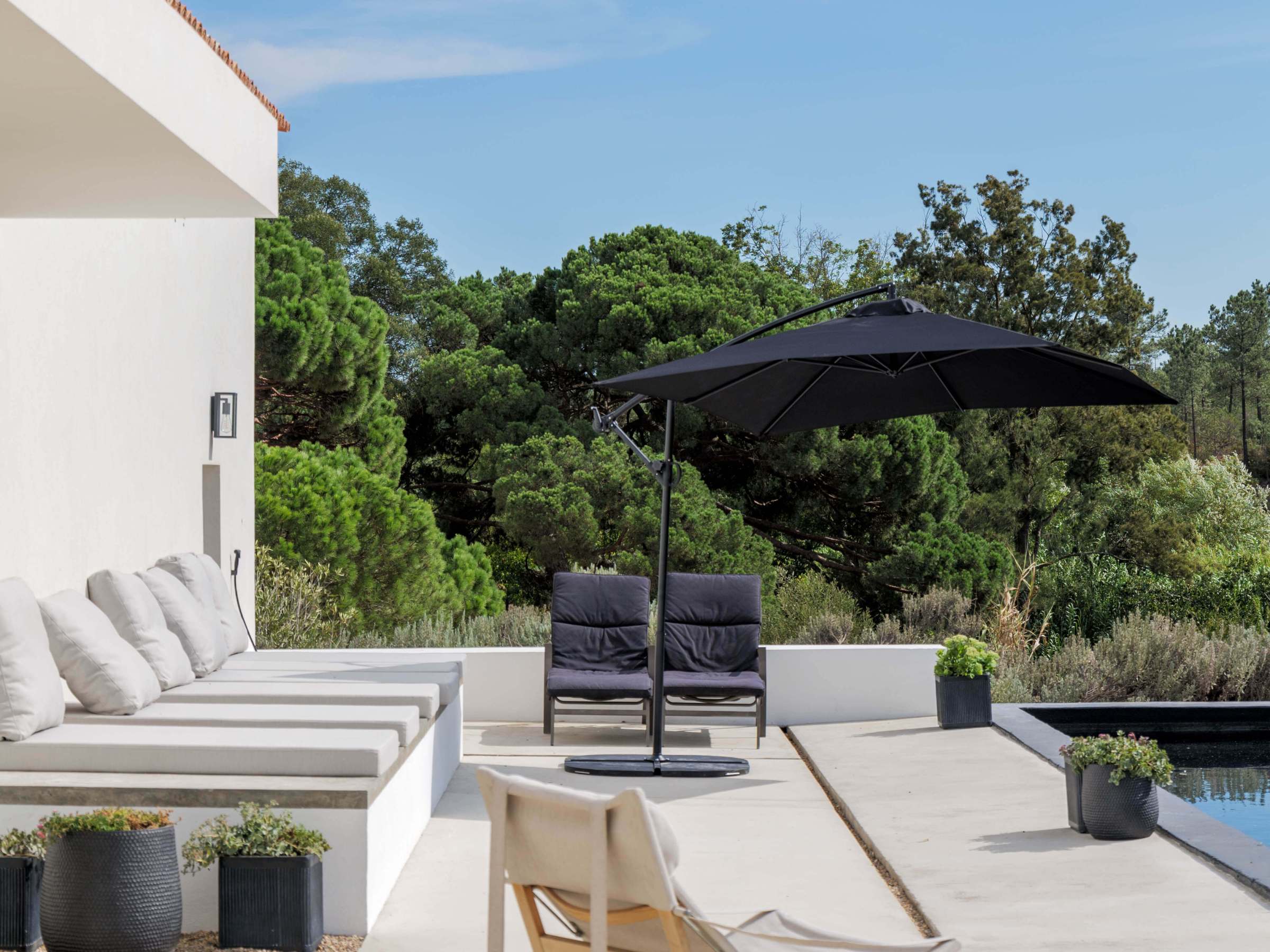 Modern patio with lounge chairs, umbrella, and greenery beside a pool under a blue sky.