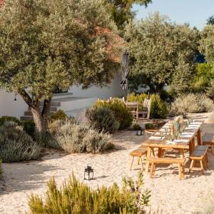 Outdoor dining area with a long wooden table and benches in a garden with trees and shrubs.